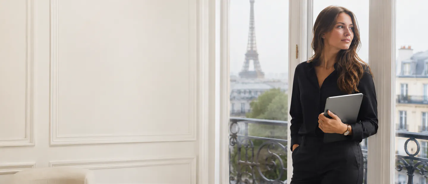 Formatrice indépendante debout, ordinateur portable à la main, devant une fenêtre ouverte sur les toits de Paris
